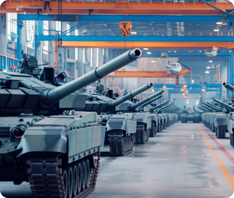 A row of military tanks lined up inside a large, industrial warehouse with cranes and other machinery visible in the background.